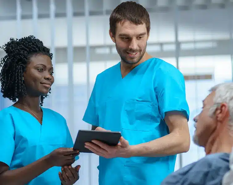 Home 9 Stem Cells Dos profesionales sanitarios, vestidos con uniformes azules, colaboran revisando el historial médico digital en una tableta mientras atienden a un paciente hospitalizado.